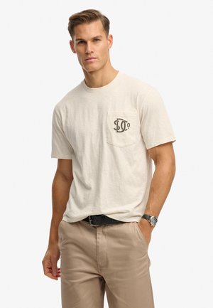 Beige Baumwoll-T-Shirt mit Rundhalsausschnitt, kurzen Ärmeln und einer linken Brusttasche mit dunklem Grafikdesign. Kombiniert mit hellbraunen Hosen.