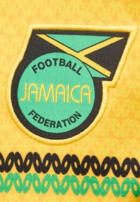 Μπατς με το λογότυπο της Ομοσπονδίας Ποδοσφαίρου της Τζαμάικας με σχέδιο σημαίας σε πράσινο, κίτρινο και μαύρο χρώμα πάνω σε κίτρινο ύφασμα με μαύρους και πράσινους διακοσμητικούς βρόχους.