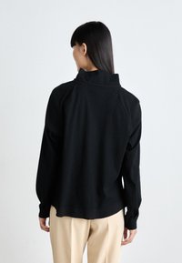 Black pullover top with a high collar, smooth texture, and long sleeves. Worn with beige pants, featuring a relaxed fit and minimal seams.