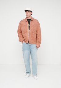 Πορτοκαλί μπουφάν με καπιτονέ σχέδιο και κουμπιά τύπου πατιλέτα, μαύρο γραφικό μπλουζάκι από μέσα, ανοιχτό μπλε φαρδιά τζιν και λευκά αθλητικά παπούτσια. Άσπρο καπέλο στο κεφάλι.