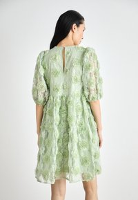 Robe en dentelle verte avec des appliqués floraux 3D, des manches bouffantes courtes, une ouverture en clé de dos et une silhouette de jupe évasée.