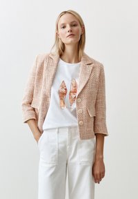 Veste en tweed pêche avec un col cranté et trois boutons, portée sur un t-shirt blanc affichant un graphique de chaussure. Associée à un pantalon blanc à jambes larges.