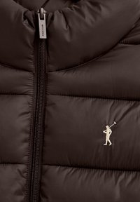 Närbild av mörkbrun quiltad jacka med vertikal dragkedja och vit broderad logga av en polospelare på bröstet.