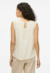 Blusa sin mangas de color crema con un cierre en la parte posterior tipo ojal y lazo, con un detalle de encaje texturizado en los hombros. Tejido suave y fluidos.
