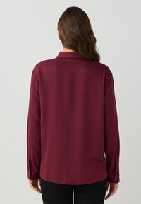 Femme avec de longs cheveux bruns portant un blouse bordeaux à manches longues et un pantalon noir, vue de dos sur un fond uni.