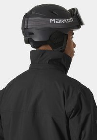 Chaqueta de esquí negra con un cuello alto, que presenta una tela texturizada. La persona lleva un casco negro con una correa ajustable gris.