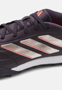 Botas de fútbol sintéticas negras con rayas texturizadas plateadas y rojas, que cuentan con cordones negros y una suela blanca para tracción.