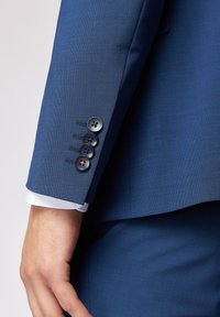 Maniche di una giacca blu con finitura testurizzata, con quattro bottoni scuri e un polsino bianco che spunta sotto la manica.