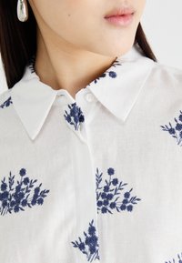 White linen shirt featuring blue botanical embroidery, a classic collar, and a button closure at the neck, with a smooth texture.