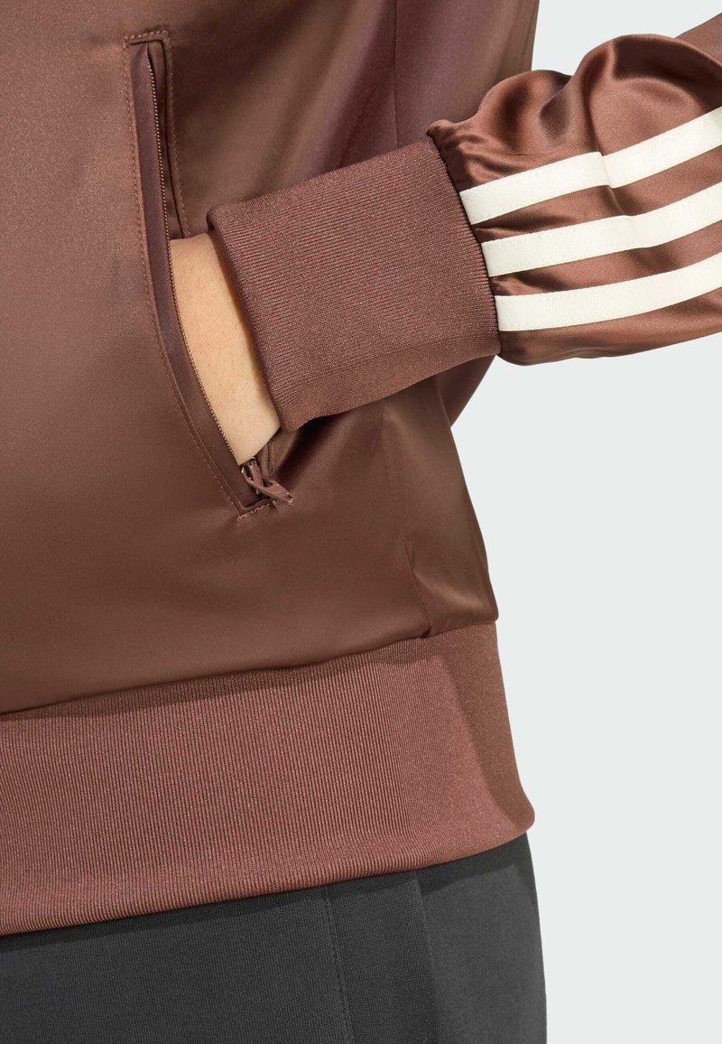 Brown satin jacket featuring a zippered pocket, ribbed cuffs, and three white stripes accent on the sleeve, with a smooth texture.