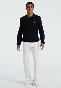 Mann steht da und trägt einen dunkelblauen Strickpullover mit Kragen und Knöpfen, weiße Hosen und weiße Sneaker vor einem schlichten hellen Hintergrund.