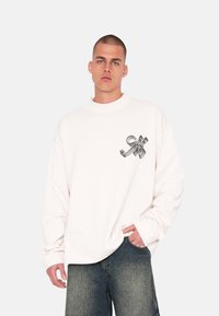 Light-colored, oversized sweatshirt with a crew neck and a metallic logo print on the chest. Crafted from a soft, textured fabric.