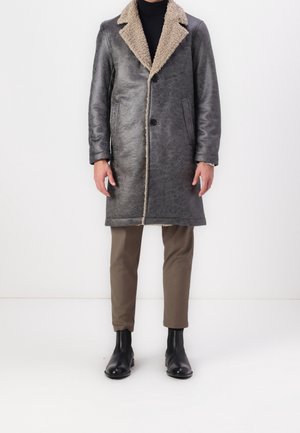 Mężczyzna w szarym płaszczu z owczej skóry sięgającym do kolan z beżowym futerkiem w środku, brązowych spodniach, czarnym golfie i czarnych skórzanych butach.