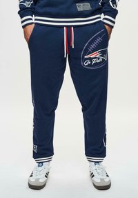 Navy-coloured collegehousut, valmistettu puuvilla-sekoitteesta, valkoisilla ja punaisilla kiristyshoitoilla, joissa on jalkapallograafika ja "Go Pats" -teksti, ribattu helmassa ja logo-tarrat.