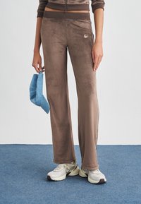 Άτομο που φοράει καφέ βελουτέ παντελόνι Nike, μπεζ παπούτσια Nike, κρατώντας μπλε τζιν καπέλο δίπλα του, στέκεται σε μπλε χαλί.