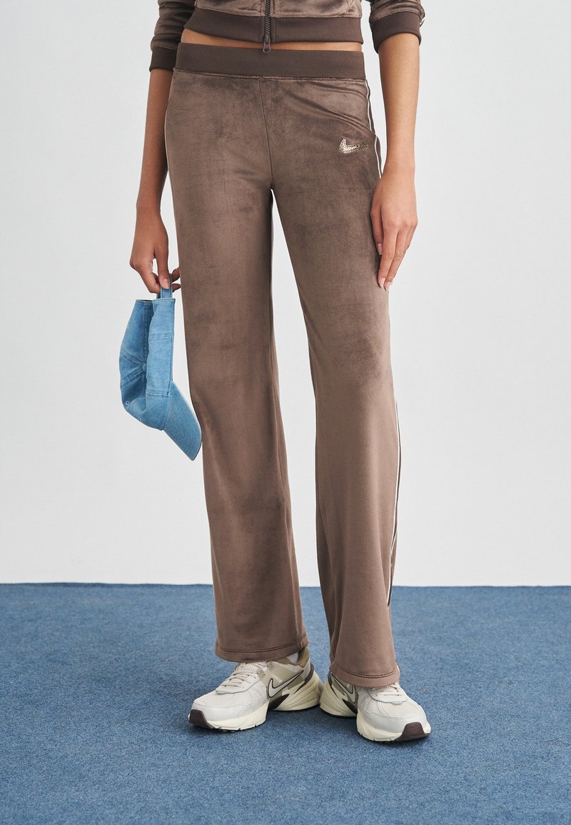 Άτομο που φοράει καφέ βελουτέ παντελόνι Nike, μπεζ παπούτσια Nike, κρατώντας μπλε τζιν καπέλο δίπλα του, στέκεται σε μπλε χαλί.