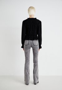 Personne aux cheveux blonds courts se tient face à un mur blanc, portant un haut noir à manches longues et un jean flare gris délavé.