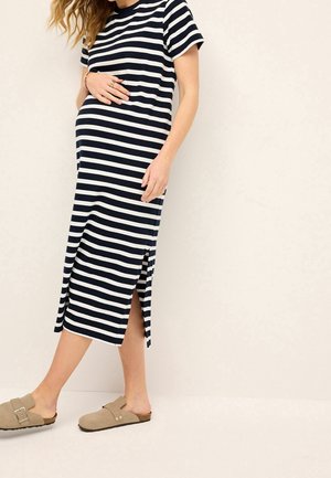 Femme enceinte portant une robe midi rayée noire et blanche avec une fente latérale et des chaussures beiges à enfiler, debout devant un fond uni.