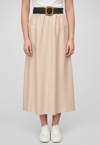 Longue jupe beige en tissu texturé, avec une taille froncée et une ceinture tissée noire ornée d'une grande boucle en bois rectangulaire. Associée à des baskets blanches.
