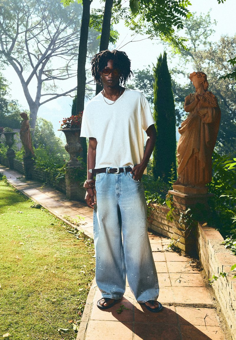 Man draagt oversized jeans, wit t-shirt, zonnebril en sandalen, staand op een tuinpad vlakbij klassieke beelden in daglicht.