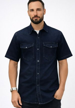 Homme barbu portant une chemise en jean bleu foncé à manches courtes avec poches poitrine boutonnées, un sous-t-shirt blanc et une montre-bracelet, debout devant un fond blanc.
