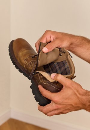 Hände halten einen braunen Lederstiefel mit Reißverschluss, der das karierte Innenfutter und das Größenetikett im Inneren zeigt.