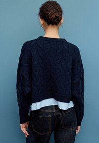 Maglione blu navy a trecce con design corto e orlo a coste, indossato sopra una camicia azzurro chiaro, abbinato a jeans denim scuro.