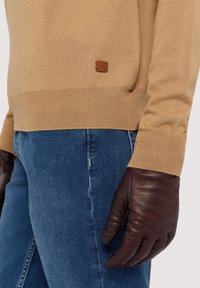 Beige stickad tröja med ribbade muddar, kombinerad med mörkbruna skinnhandskar. Märkbar skinnlogotyp på tröjans vänstra sida. Denimjeans syns.