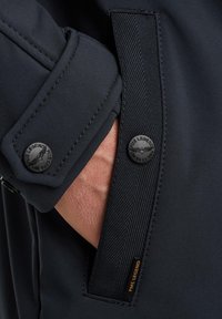 Detalj mornarskog sakoa koji prikazuje bliži pogled na manžet s crnim dugmadi i teksturiranim materijalom. Logo je otisnut na dugmetu.