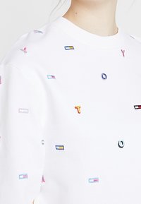 Vit sweatshirt med flerfärgad broderad geometrisk form och bokstäver. Rund halsringning med ribbad detalj vid ärmslut och nederkant.