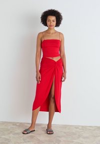 Conjunto de dos piezas rojo que incluye un top ajustado con tirantes finos y una falda hasta la rodilla con un detalle retorcido y una abertura alta. Textura de tela suave.