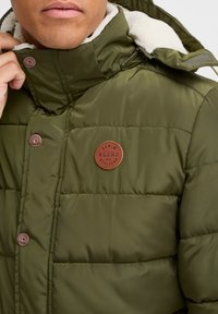Polstret olivengrøn jakke med høj krave, der har en cremet faux shearling foring og et brunt læderlogo. Bronze trykknapper synlige.