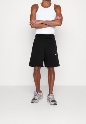 Mies, jolla on päällään valkoinen hihaton toppi, mustat kuminauhavyötärölliset shortsit, joissa on sivutaskut, ja harmaat juoksukengät, seisoo käsivarret ristissä.