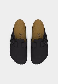 Ζευγάρι μαύρων παντόφλων slip-on με λουριά με αγκράφες, ταμπα πέλματα και σημάδια μεγέθους ορατά μέσα σε κάθε παπούτσι σε λευκό φόντο.