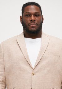 Blazer de lino beige con un solo botón, solapas de muesca y detalle de bolsillo lateral, llevado sobre una camiseta blanca contra un fondo neutro.