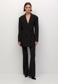 Frau steht vor einem schlichten Hintergrund und trägt einen schwarzen oversized Blazer sowie passende auslaufende Hosen mit spitzen schwarzen Absätzen.