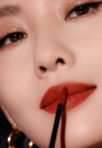 Applicateur de rouge à lèvres liquide tenu près de lèvres rouges profondes, avec une texture matte lisse. Un maquillage des yeux doux et une peau éclatante mettent en valeur les caractéristiques.
