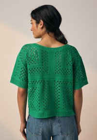 Tumejuustega naine, kellel on madal pats, seljas roheline heegeldatud lühikeste varrukatega top ja sinised teksad, seisab ühetaustalise heleda tausta ees.