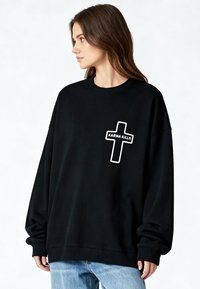 Femme aux cheveux bruns portant un sweat-shirt noir surdimensionné avec une croix blanche et le texte « KARMA KILLS », assorti d'un jean bleu clair, regardant vers la gauche.