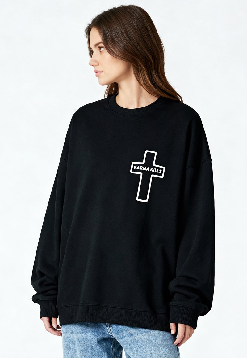 Femme aux cheveux bruns portant un sweat-shirt noir surdimensionné avec une croix blanche et le texte « KARMA KILLS », assorti d'un jean bleu clair, regardant vers la gauche.