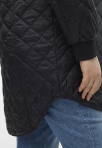 Chaqueta acolchada negra con dobladillo redondeado, que presenta una superficie texturizada y puños elásticos, combinada con vaqueros de mezclilla azul.
