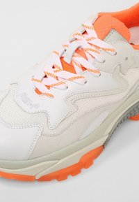 Baskets de sport blanches et orange avec des lacets blancs et orange à motifs, des panneaux en mesh et en cuir, et une semelle texturée orange.