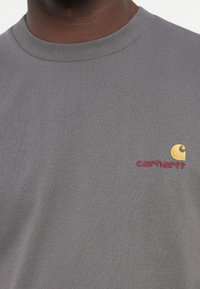 Gray cotton t-shirt with a ribbed crew neck. Features a small yellow and pink embroidered logo on the left chest area.