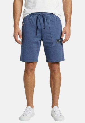 Homme portant un short bleu à cordon avec des poches avant et un patch logo sur la poche droite, accompagné de baskets blanches.