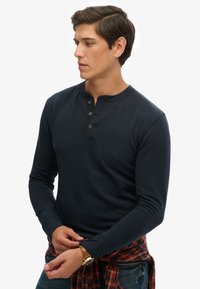 Camisa henley de manga larga en azul marino con un cuello redondeado, tapeta de tres botones y mangas con puños. Llevada con una camisa de cuadros atada a la cintura.