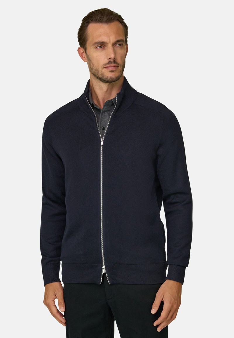 Homme aux cheveux bruns courts portant un pull zippé bleu marine foncé par-dessus une chemise grise à col, debout devant un fond clair uni.