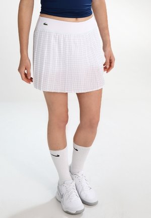 Frau trägt weißen plissierten Tennisrock mit Rautenmuster, weiße Nike-Sneaker und weiße Nike-Socken, steht vor weißem Hintergrund.