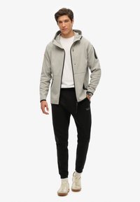 Sudadera con cremallera de color gris claro, con capucha y bolsillos, combinada con pantalones jogger negros y zapatillas beige. Tejido suave con acentos en contraste.