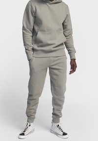 Felpa grigia in cotone con tasca a marsupio e pantaloni della tuta abbinati. Tessuto strutturato, polsini a costine e cappuccio con cordino. Indossata con scarpe bianche alte.