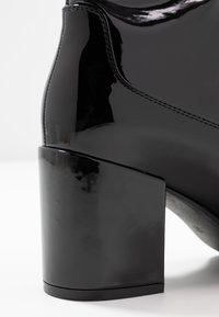 Bota de charol negro brillante con tacón de diseño cuadrado sobre un fondo blanco, mostrando detalles de costura y superficie lisa.